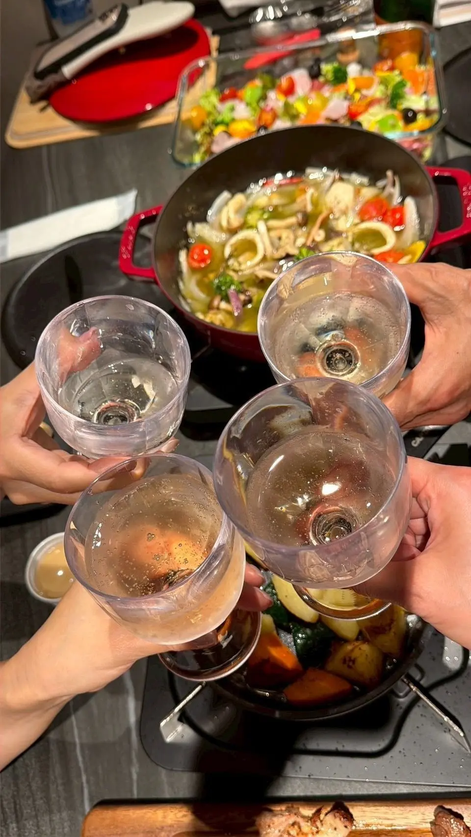 5つのワイングラスがぶつかり合って乾杯している様子。背景には赤いル・クルーゼの鍋に入った料理と、サラダが置かれたガラス容器が見える。