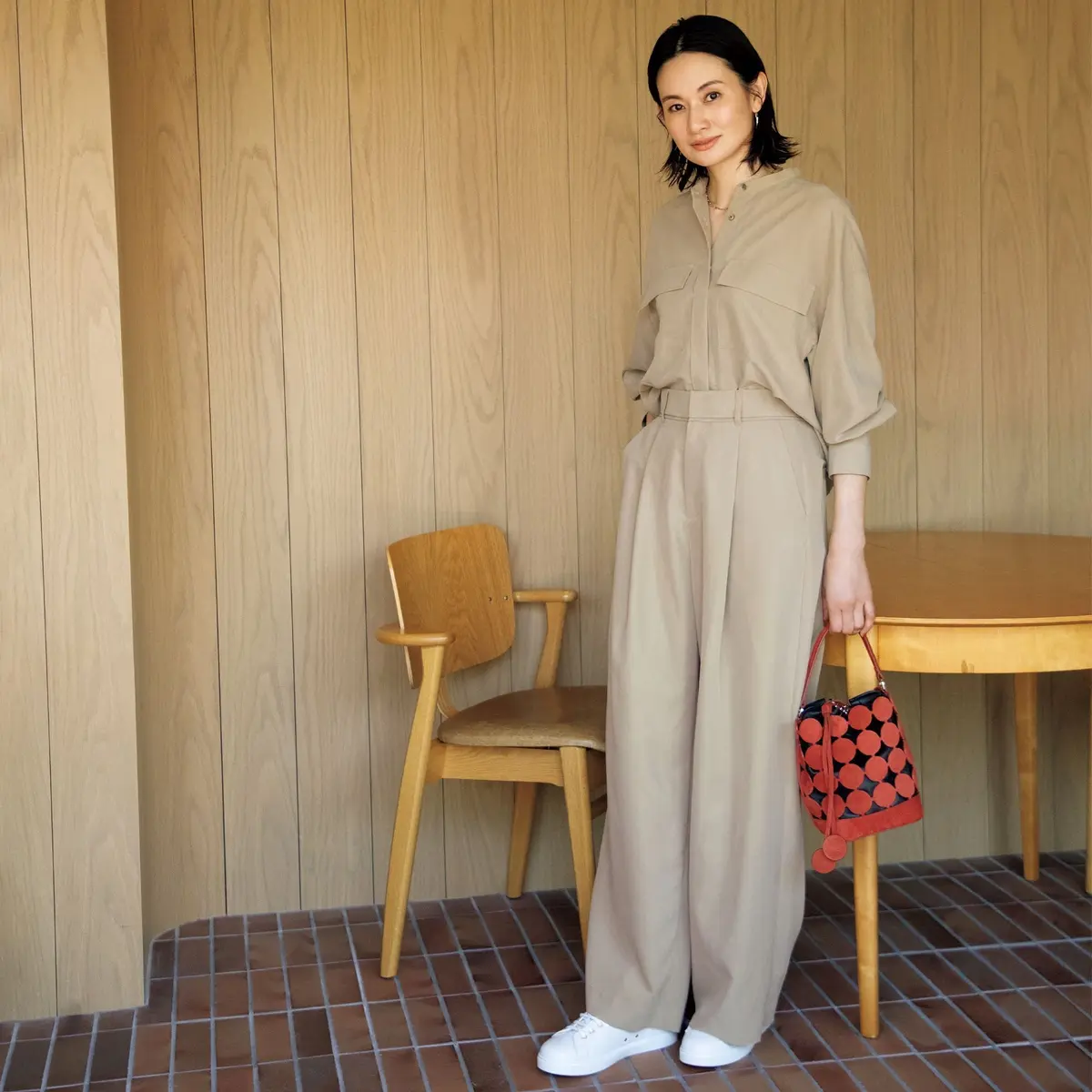 ベージュのシャツとパンツのセットアップを着た女性が、木製の壁の前に立っている。白いスニーカーを履き、黒いイヤリングを着用。手には赤と黒のドット柄のバッグを持っている。