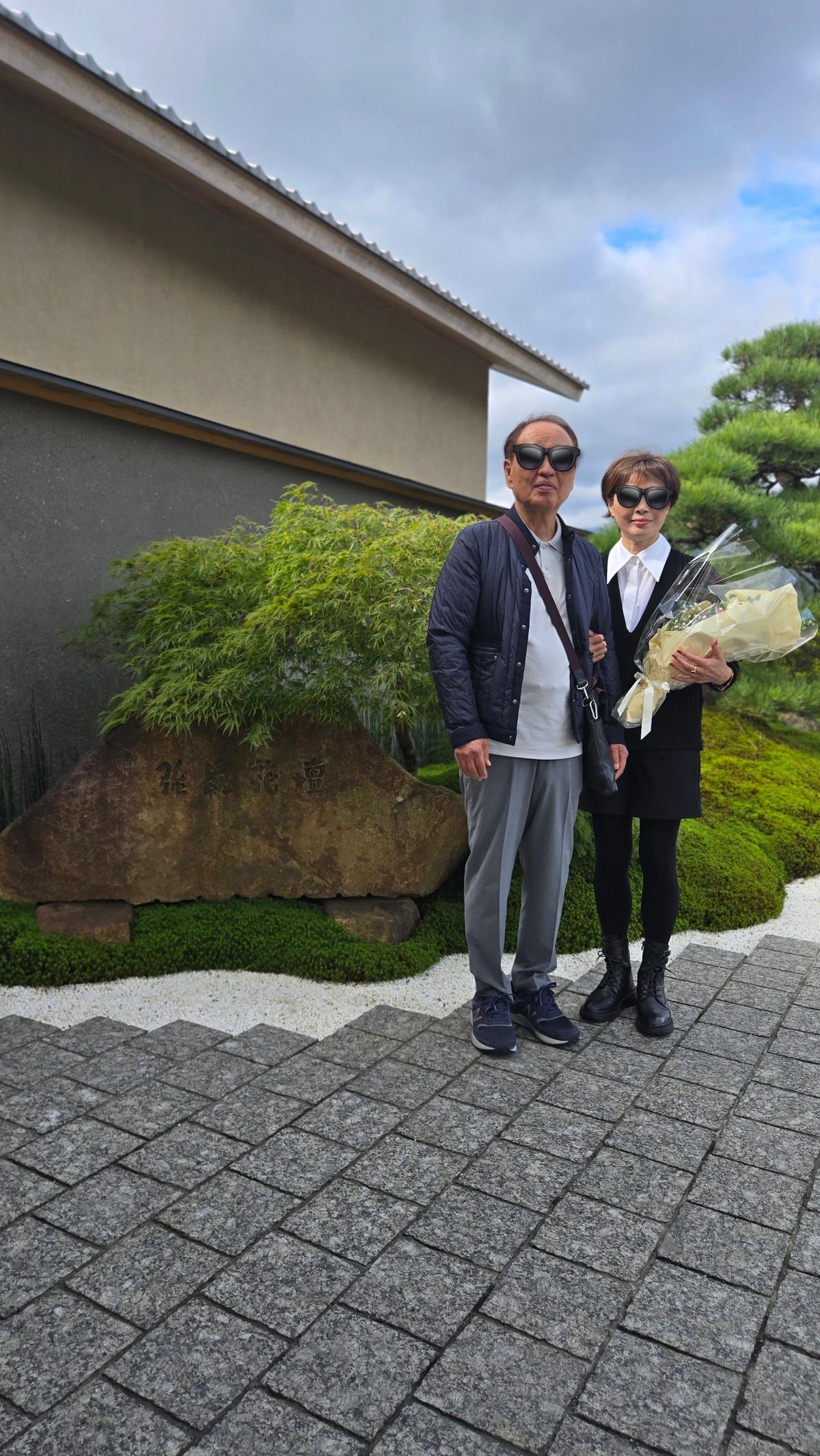 庭園を背景に、3人組が立っている写真。左から、白髪の男性、サングラスをかけた女性、黒いスーツの女性。男性はグレーのジャケットとパンツを着用。女性は白い花束を持っている。