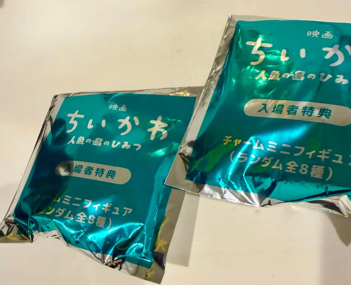 青緑色の包装紙に包まれた「ちいかわ」と書かれた映画入場者特典の文字と、チャームミニフィギュア（ランダム全8種）の記載がある。2つの包まれたミニフィギュアが斜めに配置されている。