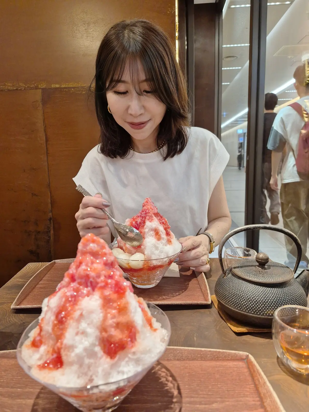 白シャツを着た女性が、木製のトレイに盛られたイチゴのかき氷をスプーンで食べている。背景にはガラス張りの空間と、茶色の木材の壁が見える。