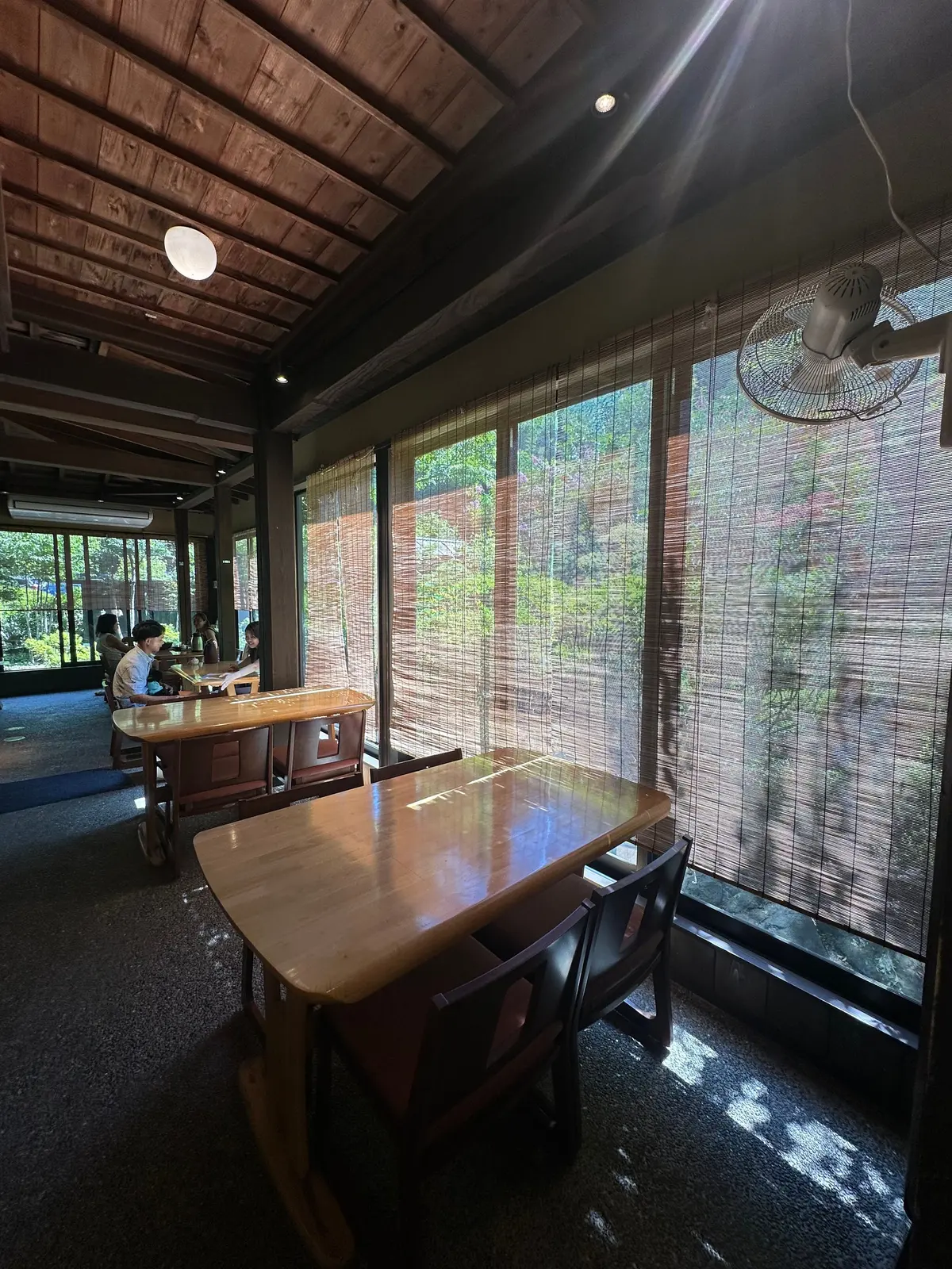 木目調の天井と大きな窓が特徴的な空間。手前に木製のテーブルと椅子、奥には人々が座っているテーブルが見える。窓には葦のブラインドが設置され、外の緑がぼんやりと見える。