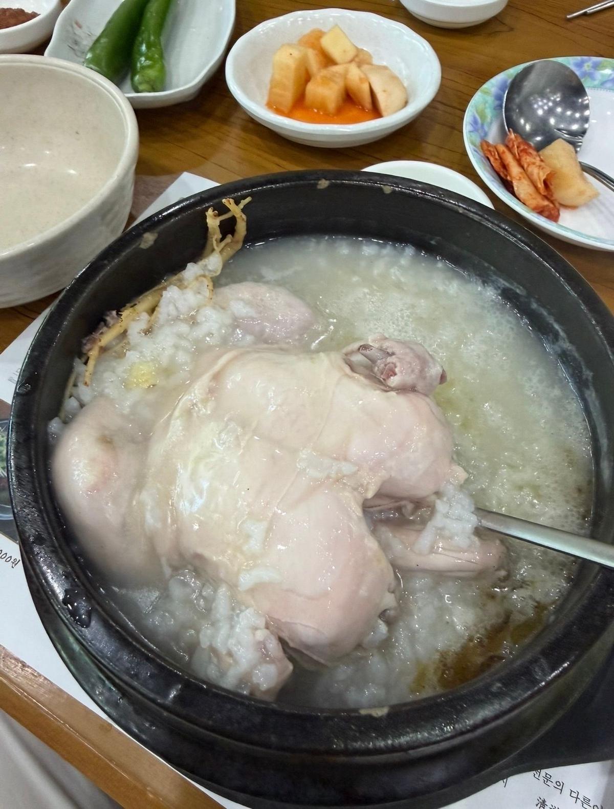 黒い鍋に入った白い乳粥に、鶏肉とキノコ、その他具材が浮かんでいる。小皿には、赤とオレンジのきんぴら、緑色の野菜、豆のペースト、魚の煮付けなどが添えられている。