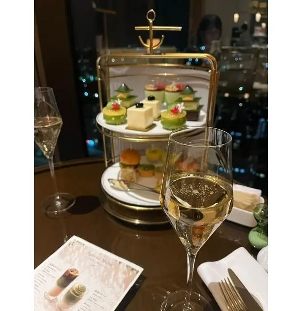 金色の2段スタンドに、様々なミニデザートが並んでいる。グラスに注がれた白ワイン、メニュー、カトラリーがテーブルに置かれている。背景には夜景が見える。