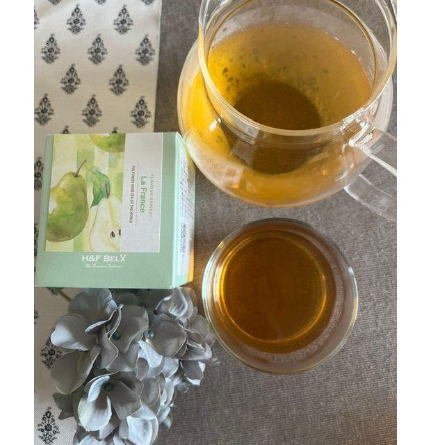 H&F BelXの「La France」ティーボックスと、2つの透明なグラスに入った黄色いお茶。背景には白い柄のテーブルクロスと青いハイビスカス。