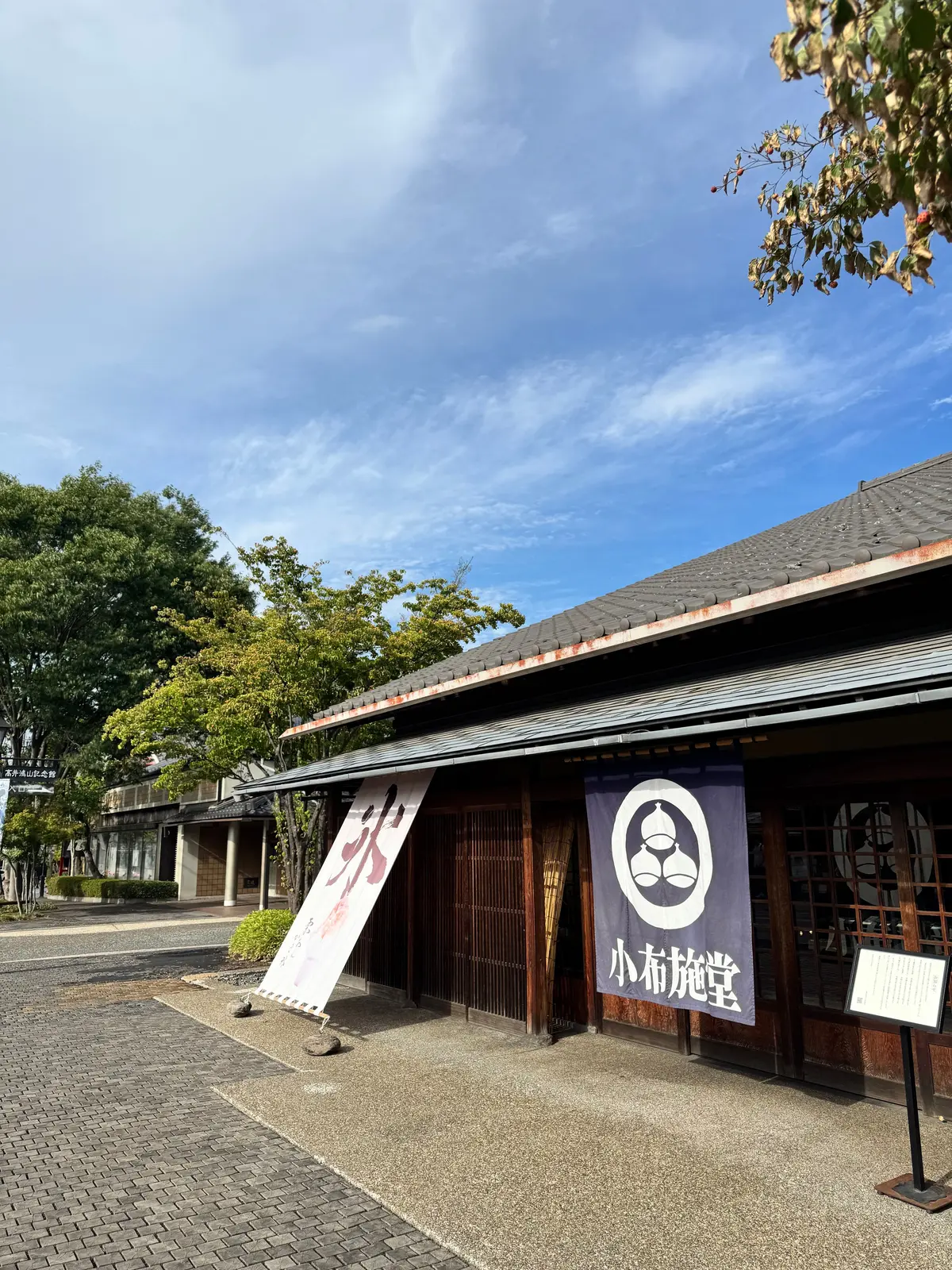小布施宿の建物。瓦屋根の茶色の木造建築で、入り口には「小布施宿」の看板が掲げられている。左には幟が立っており、青空を背景に写る。