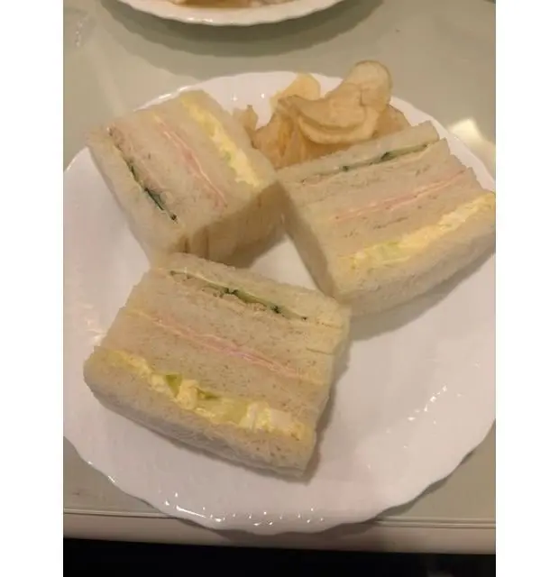 白い皿に3切れのサンドイッチとポテトチップスが置かれている。サンドイッチは食パンで挟まれ、卵、ハム、キュウリなどの具材が層状に入っている。ポテトチップスは茶色で、手前にはサンドイッチ、奥にはポテトチップスが配置されている。