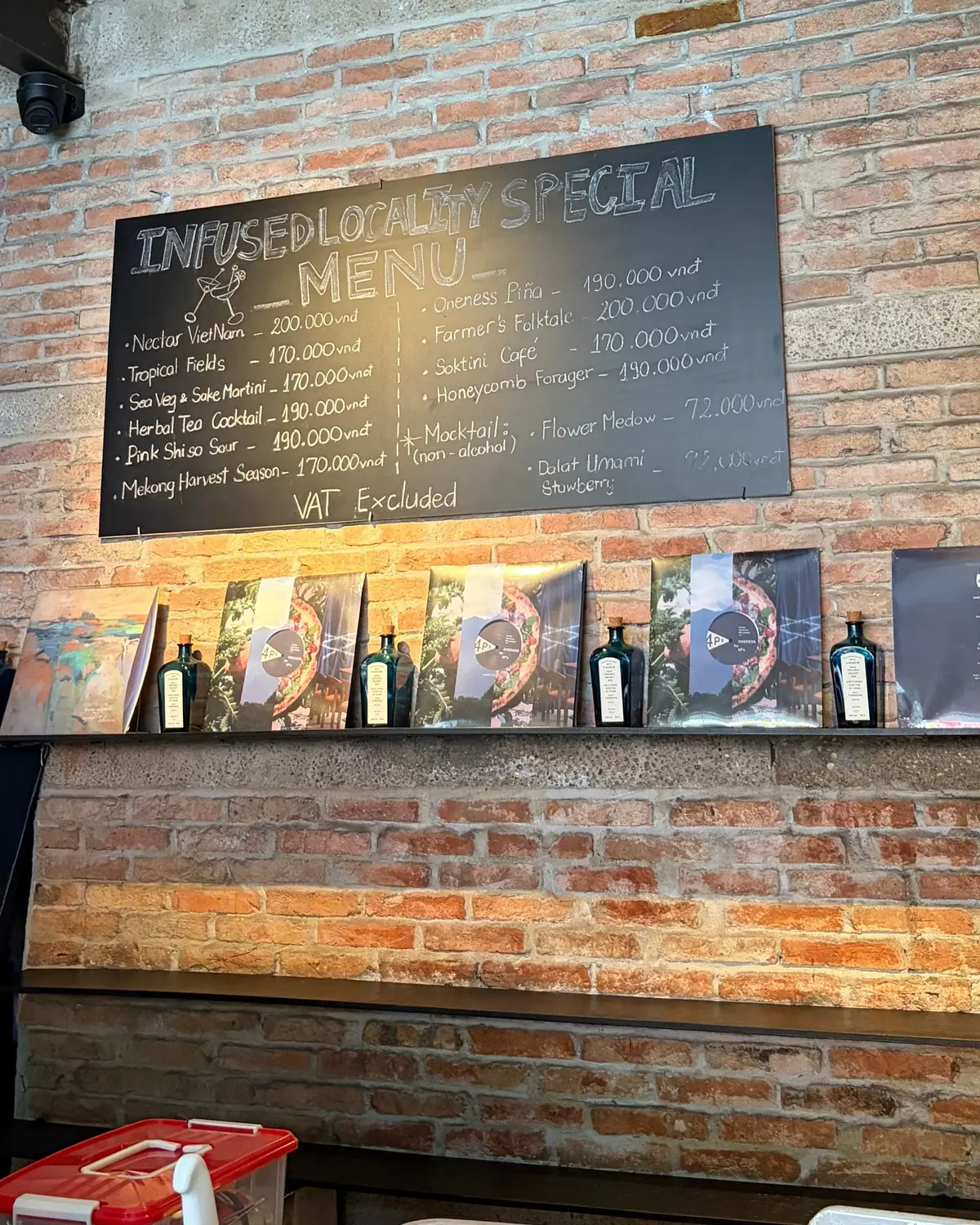 レンガの壁に「INFUSED LOCALITY SPECIAL MENU」と書かれた黒板メニューがあり、価格付きのカクテルやドリンクがリストされています。その下にはボトルとポスターが並んでいます。