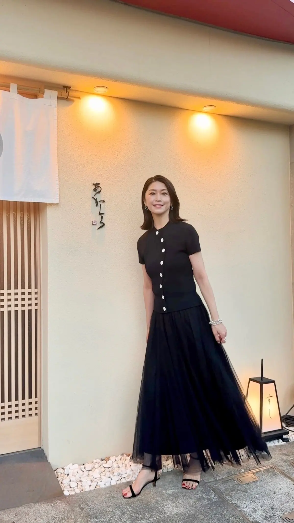 ベージュの壁に日本語の看板があり、黒のチュニックとロングスカートを着た女性が笑顔で立っている。白い布が壁に掛かり、石の敷き詰めと灯りがある。