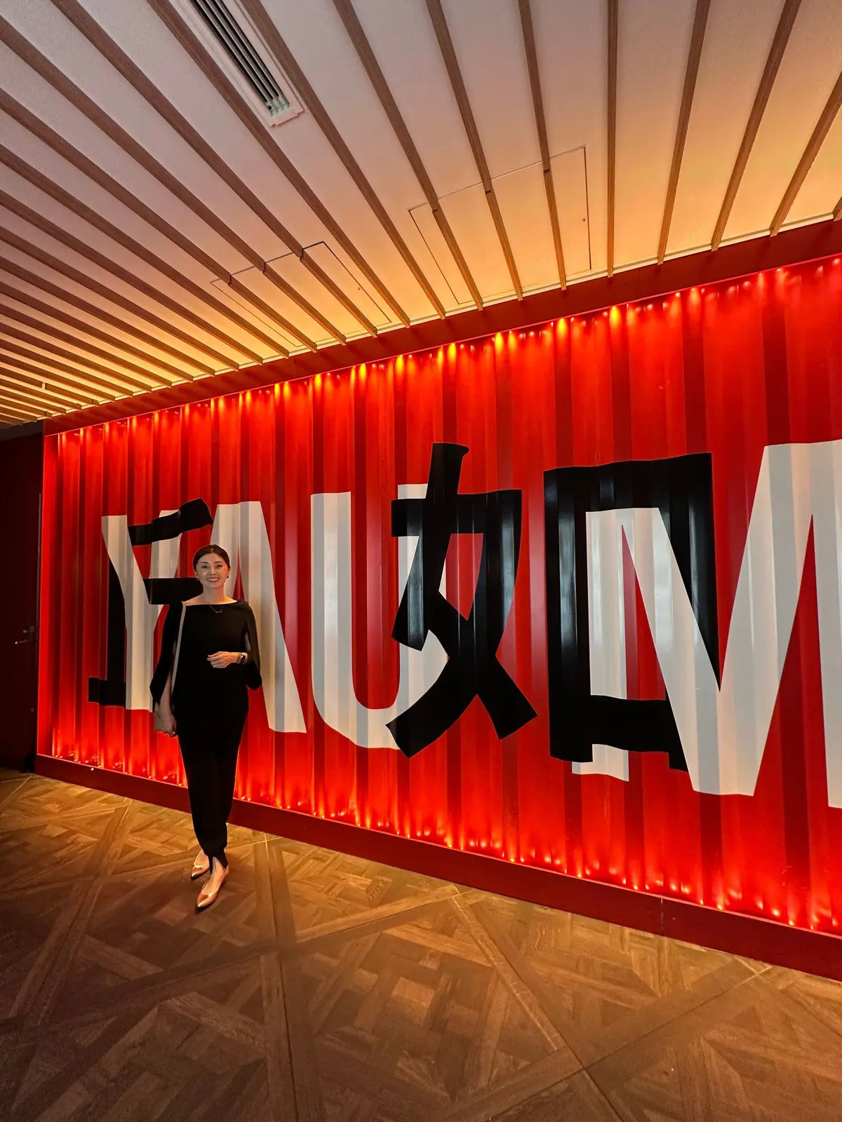 黒い服を着た女性が、赤色の縦縞の壁に大きく書かれた「FAUM」のロゴの前で立っている。天井は幾何学模様の照明で照らされている。