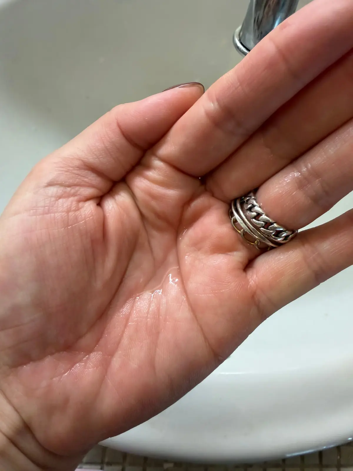 水が流れる手のひら。右手で水を待ち、シルバーのリングを着用している。背景には白い洗面台と蛇口が見える。