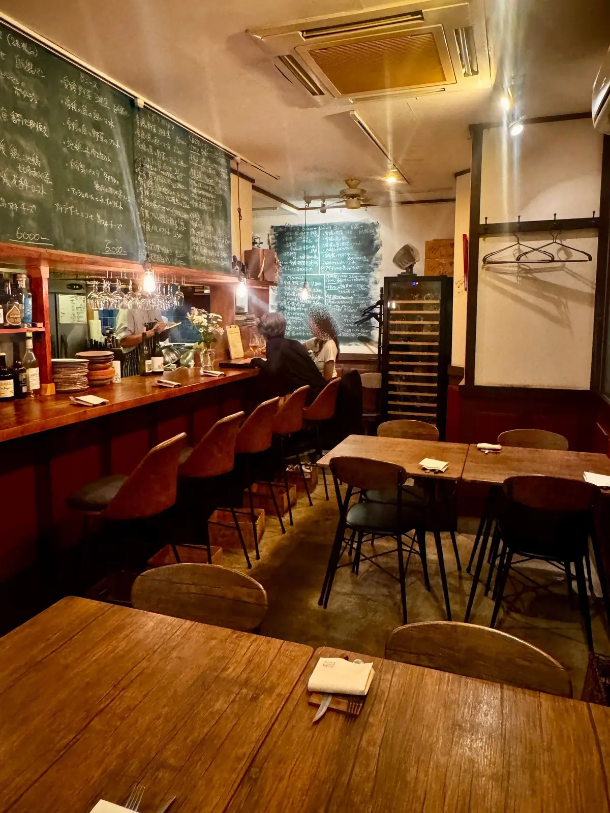 木製のカウンターとテーブルが並ぶ、落ち着いた雰囲気の飲食店内。黒板にメニューが手書きで書かれている。客席には数人の人が座り、照明が暖色系の光を放っている。