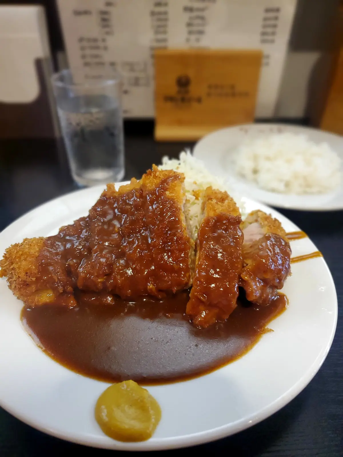 白くて丸い皿に盛り付けられた、濃い茶色のカレーと、それに浸された分厚い豚カツ。カツは斜めにカットされ、レモンとピクルスが添えられている。背景には水とご飯の盛り合わせが見える。