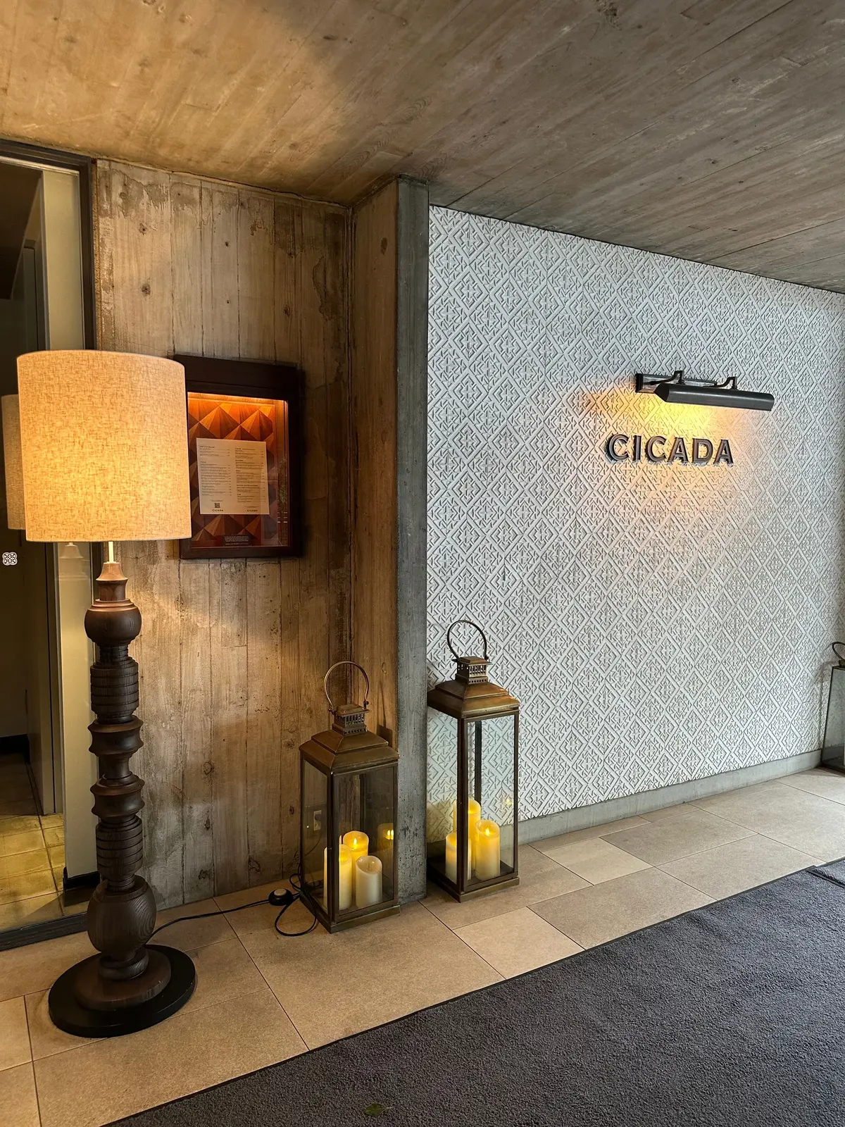 木製のフロアランプと、幾何学模様の壁に「CICADA」と書かれたサインがある空間。ランプの横には額縁に入ったメニュー、ランタン、小さな木製置物が置かれている。床はグレーのラグで覆われている。