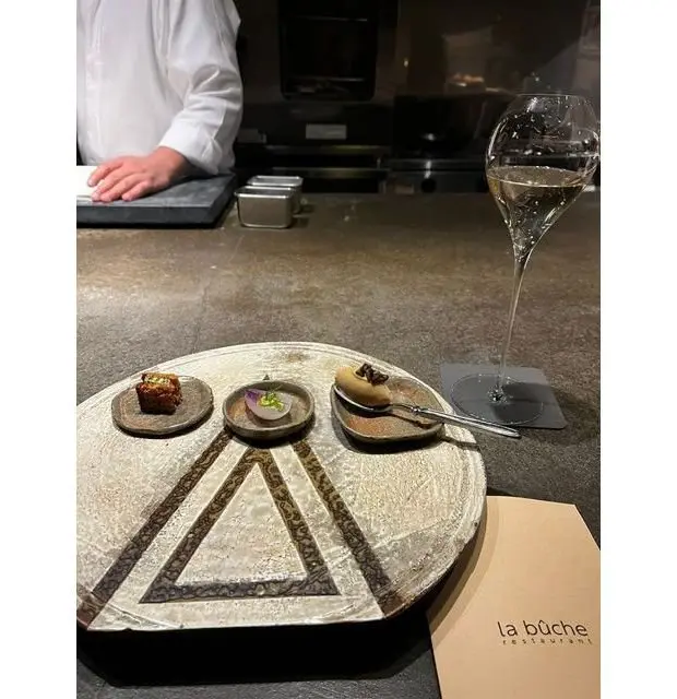 石のプレートに3つの小さな料理とワイングラスが置かれたカウンター、背景に料理人