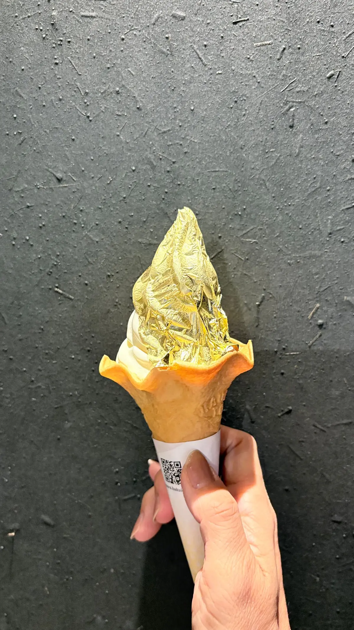 手で保持されたソフトクリーム。コーンに盛られ、表面は金箔で装飾されている。背景は黒色の壁。