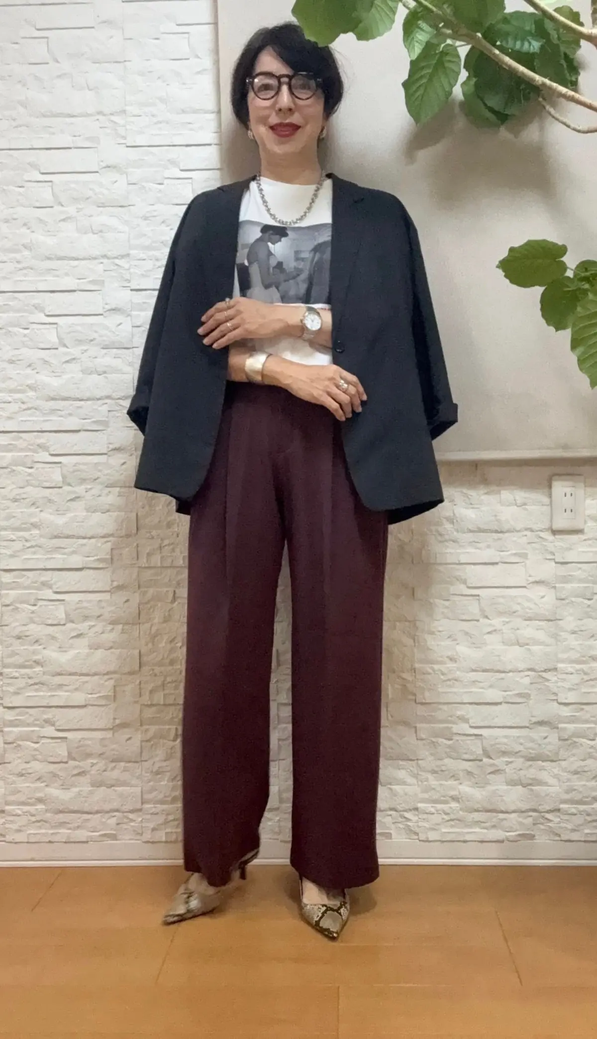 黒のジャケット、ワインレッドのワイドパンツ、白のTシャツを着用した女性。丸メガネ、パールネックレス、蛇柄のミュール。背景には白い壁と観葉植物。