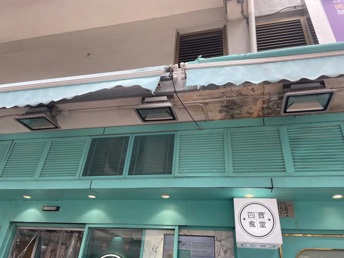 緑色の建物の店頭で、窓に緑のルーバーがあり、看板には「四喜」の文字が見える。建物の上には破れた天蓋と照明器具が取り付けられている。