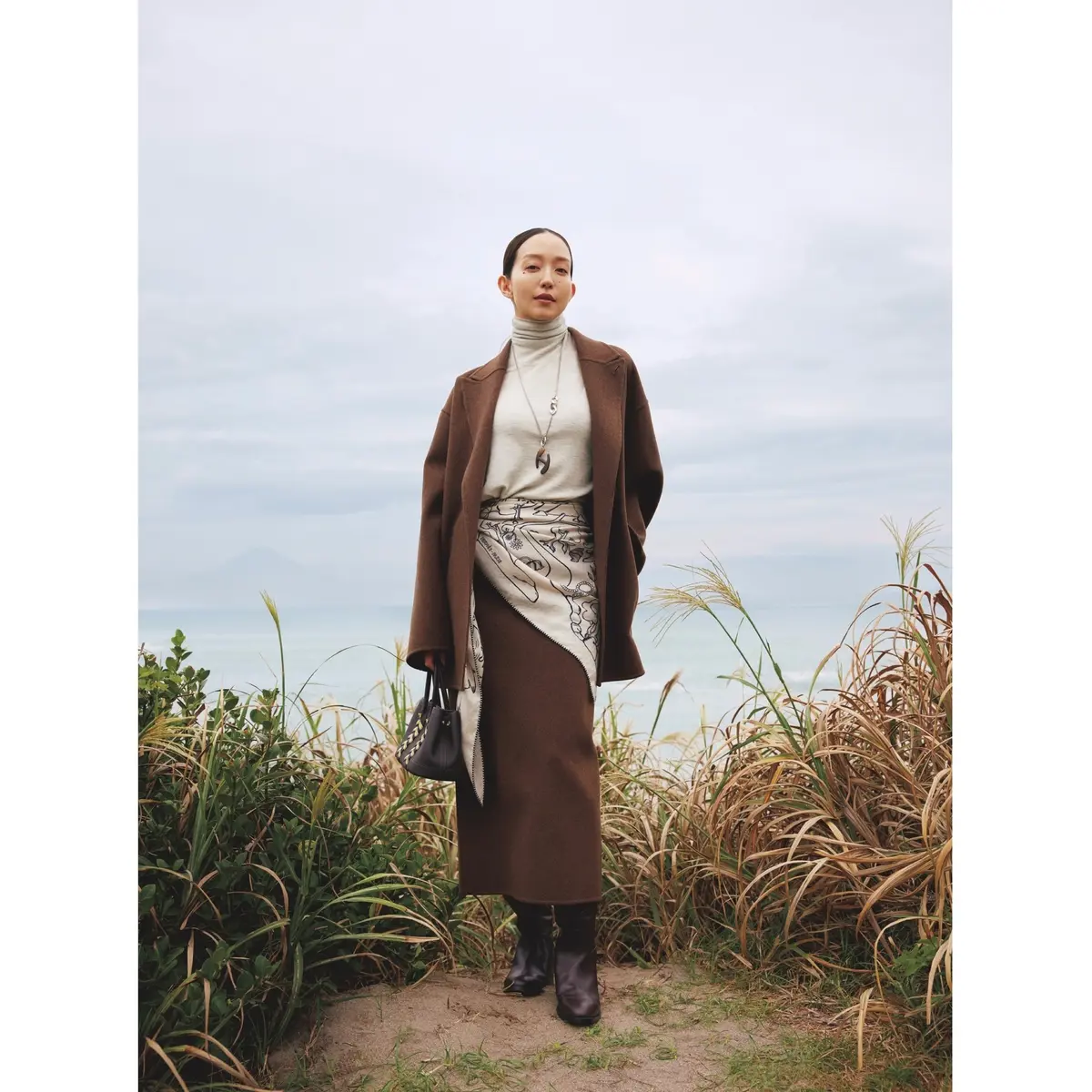 ブラウンのジャケットコートとロングスカートのセットアップに白タートルトップスを合わせ、腰にアイボリーのスカーフを巻いたコーディネート　モデル・松島花