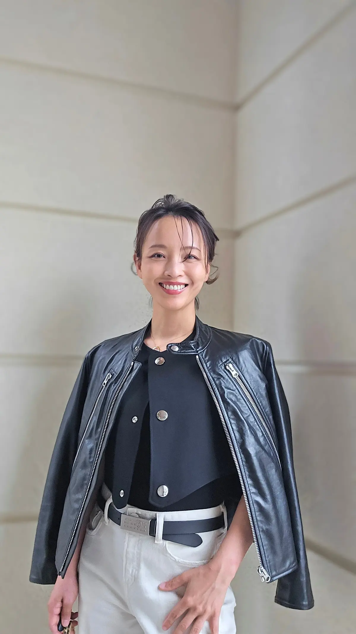 笑顔の女性が、黒のレザーJKT、黒のカットソー、白いパンツを着用。茶色のベルトを締めている。背景はベージュ色の壁。
