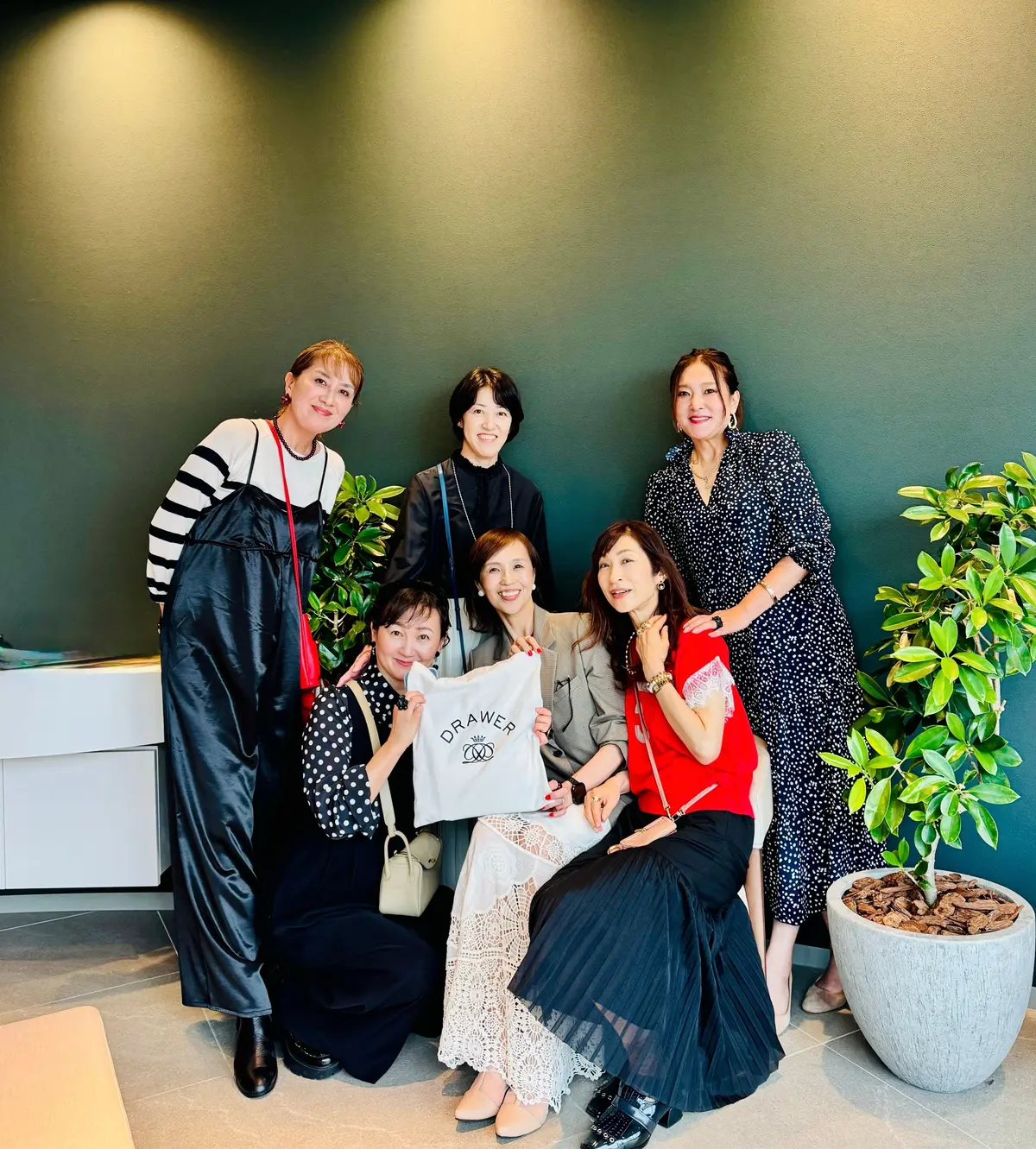 緑色の壁の前で、Drawerのロゴが入った白いバッグを持つ女性たち7人が笑顔で並んで写っている。前列中央の女性は赤いトップスに黒いスカート、後ろ列はそれぞれ異なる柄の服を着ている。