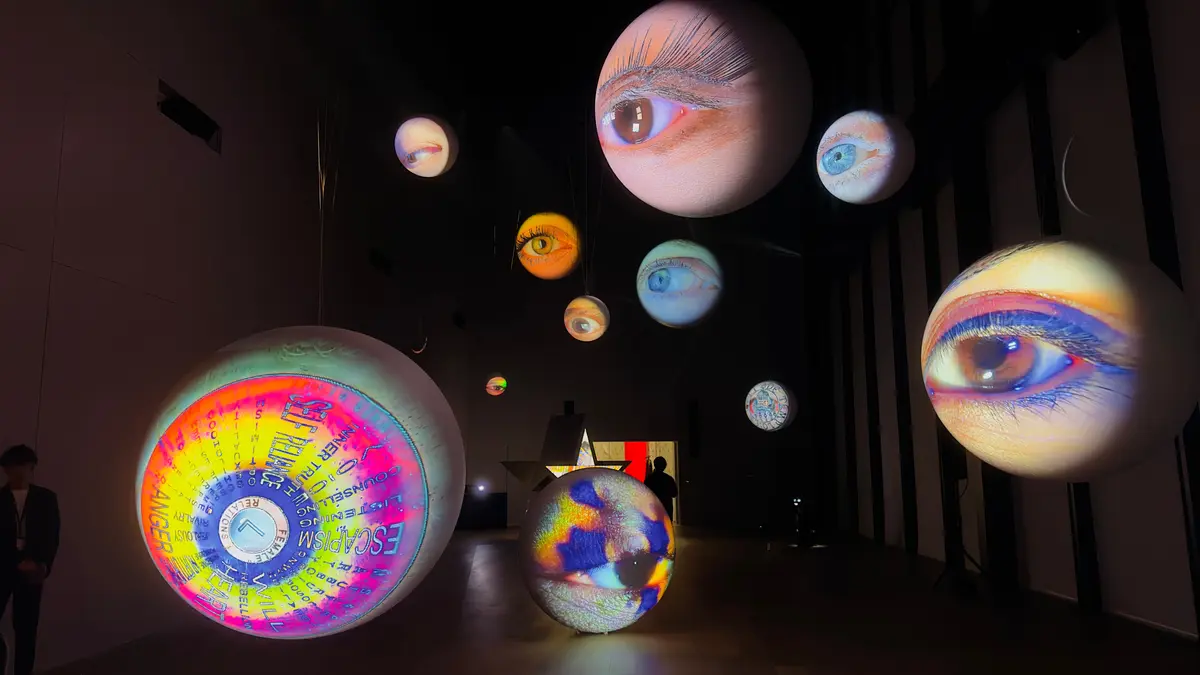 暗い空間に浮かぶ、巨大な球体型のオブジェ。それぞれに巨大な目や文字が投影され、カラフルな光を放っている。複数の球体が吊り下げられ、奥行きのある空間を演出。