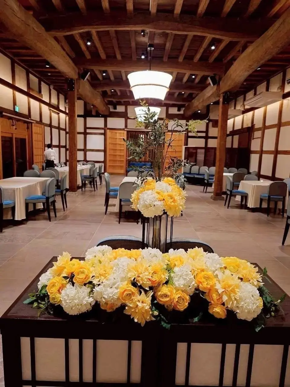 木造の梁が特徴的な広々としたレストランの内部。テーブル席が並び、中央には花のアレンジメントが置かれている。黄白色の花がテーブルや花瓶に飾られ、上品な雰囲気を演出している。