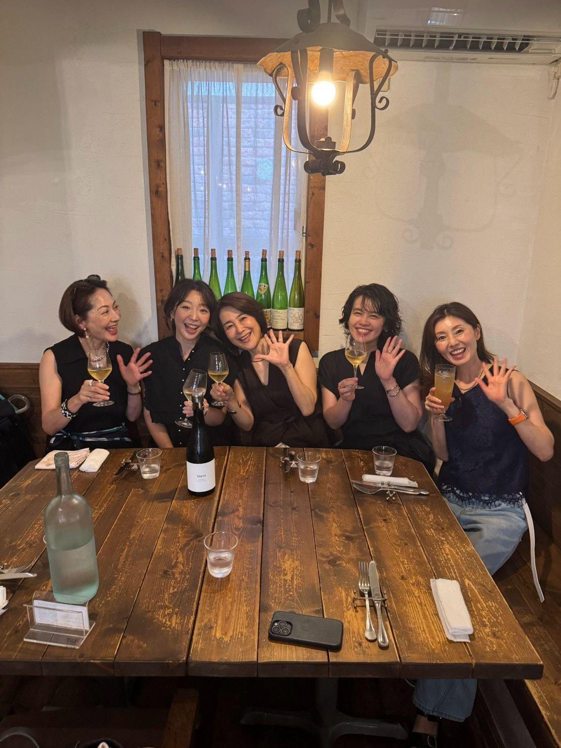 木製のテーブルを囲み、5人の女性がグラスを掲げて笑顔で写っている。背景には窓と照明器具、テーブルにはワインボトルや料理が並ぶ。