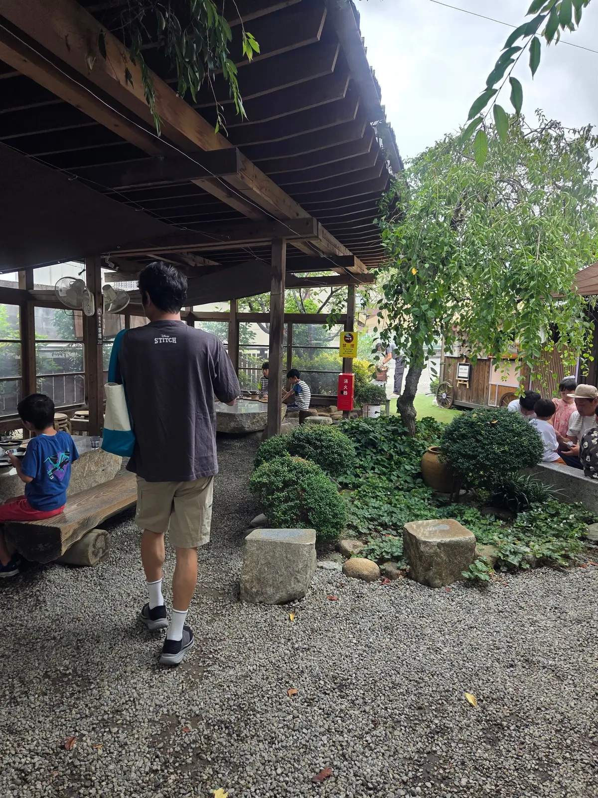 木造の屋根付き休憩スペース。男性が歩き、左には子供が座っている。手前に小石敷きがあり、緑の植栽と石段が配置されている。背景には人々や木々が見える。