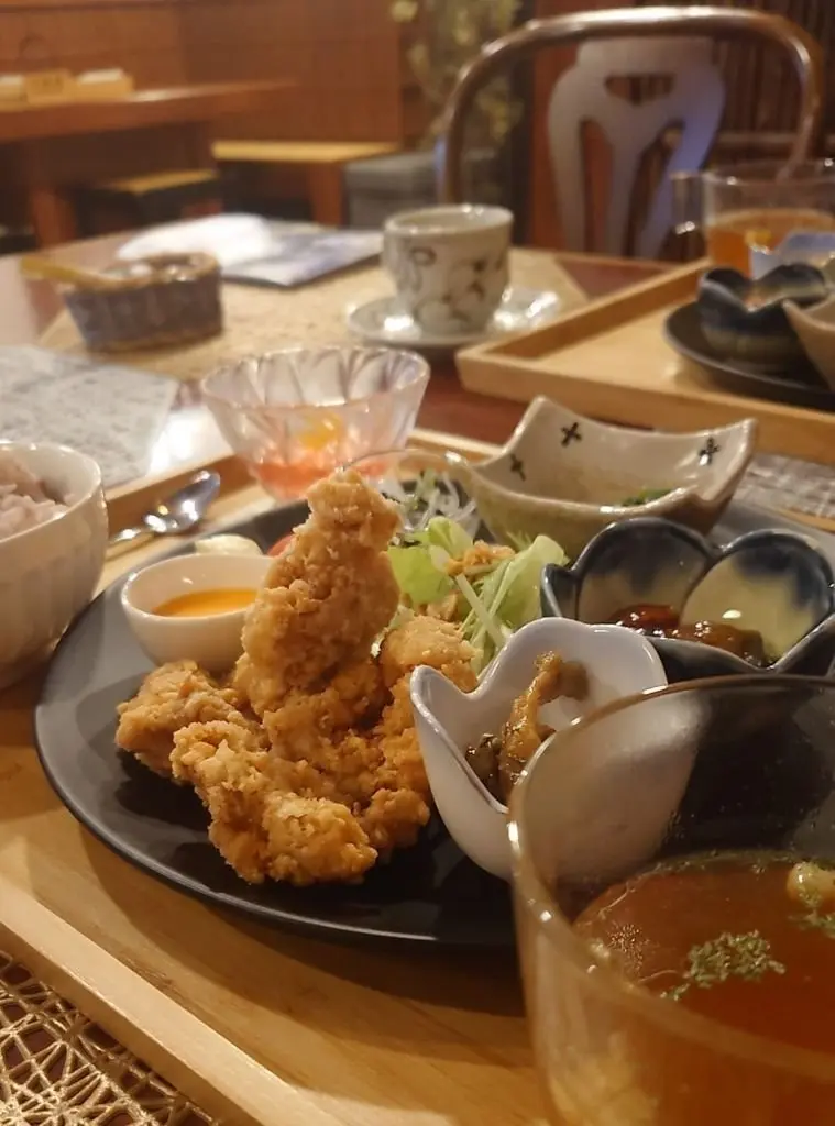 木製のトレイに盛り付けられた、鶏肉の唐揚げと様々な小皿。唐揚げは黒いお皿に盛られ、レモンソースを添えている。小皿にはご飯、漬物、魚肉ソーセージ、だし汁などが乗っている。背景にはテーブルや椅子が見える。