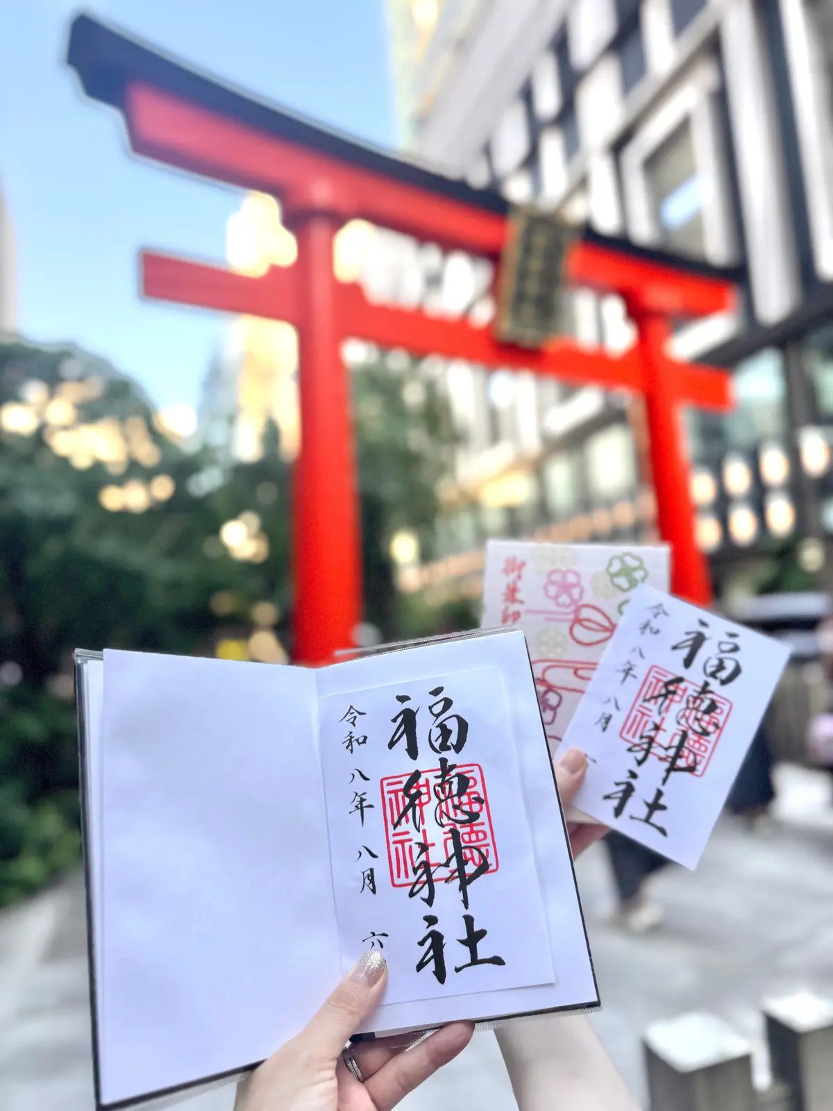 手で複数の白いおみくじを持つ女性。背景には朱色の鳥居と緑豊かな木々が見える。おみくじには黒い文字で神社名と日付が書かれている。