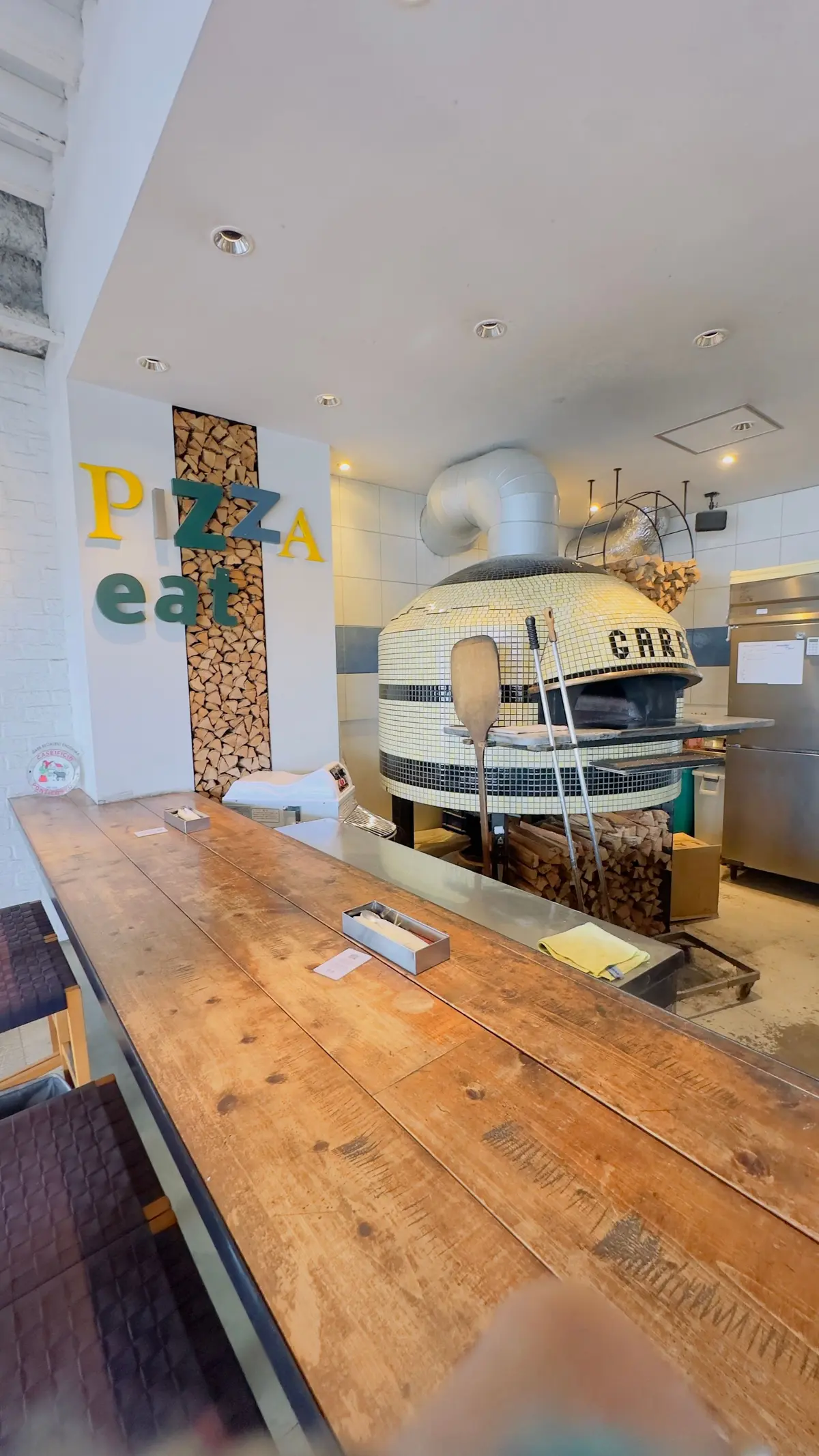 木製のカウンターと石造りのピザ窯が並ぶピザ屋の店内。壁には「PIZZA eat」の文字があり、周囲には木の薪が積まれている。