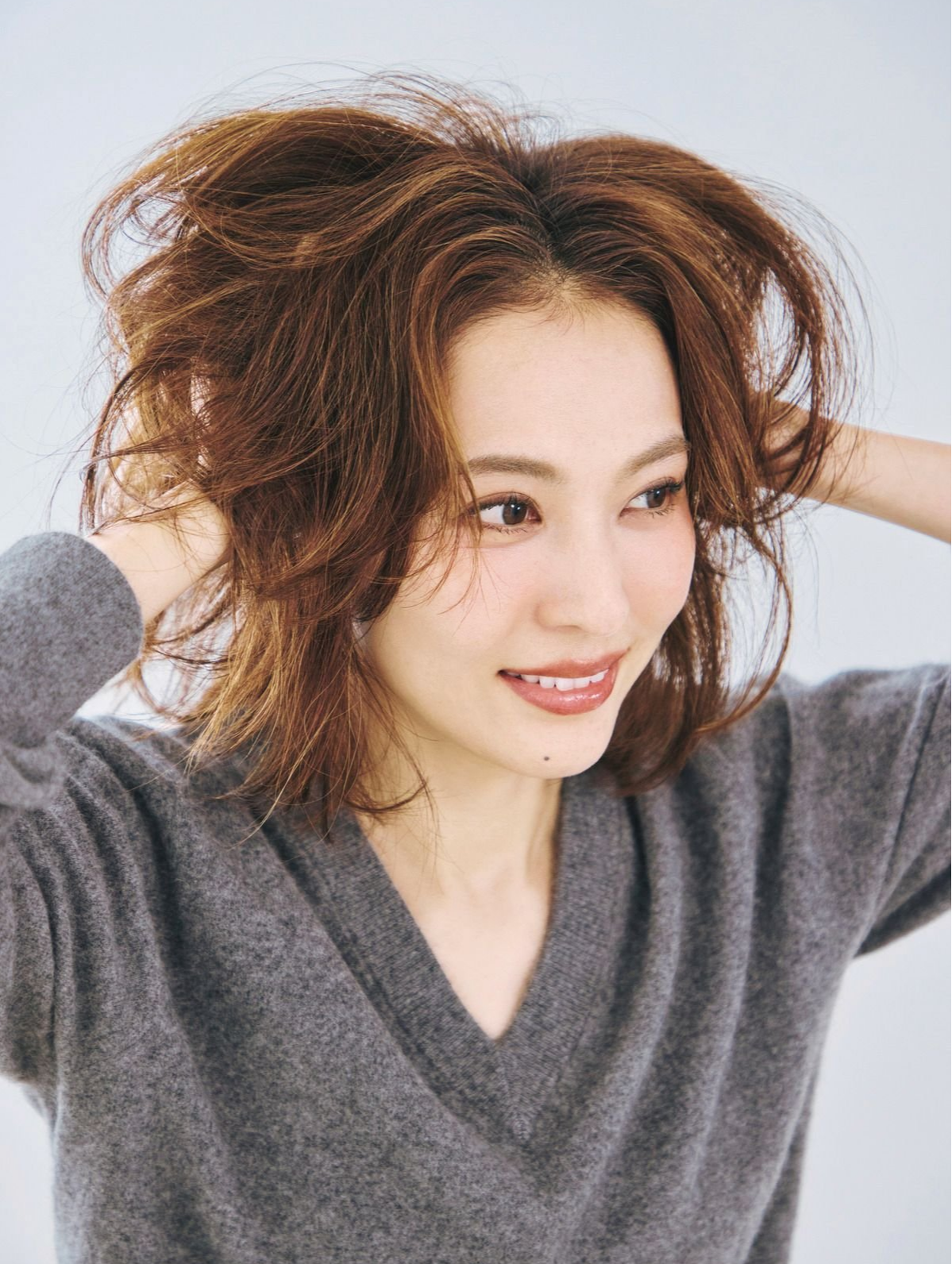 灰色のセーターを着た女性が、茶色のウェーブのかかった髪を両手で持ち上げ、微笑んでいるポートレート。背景は白。