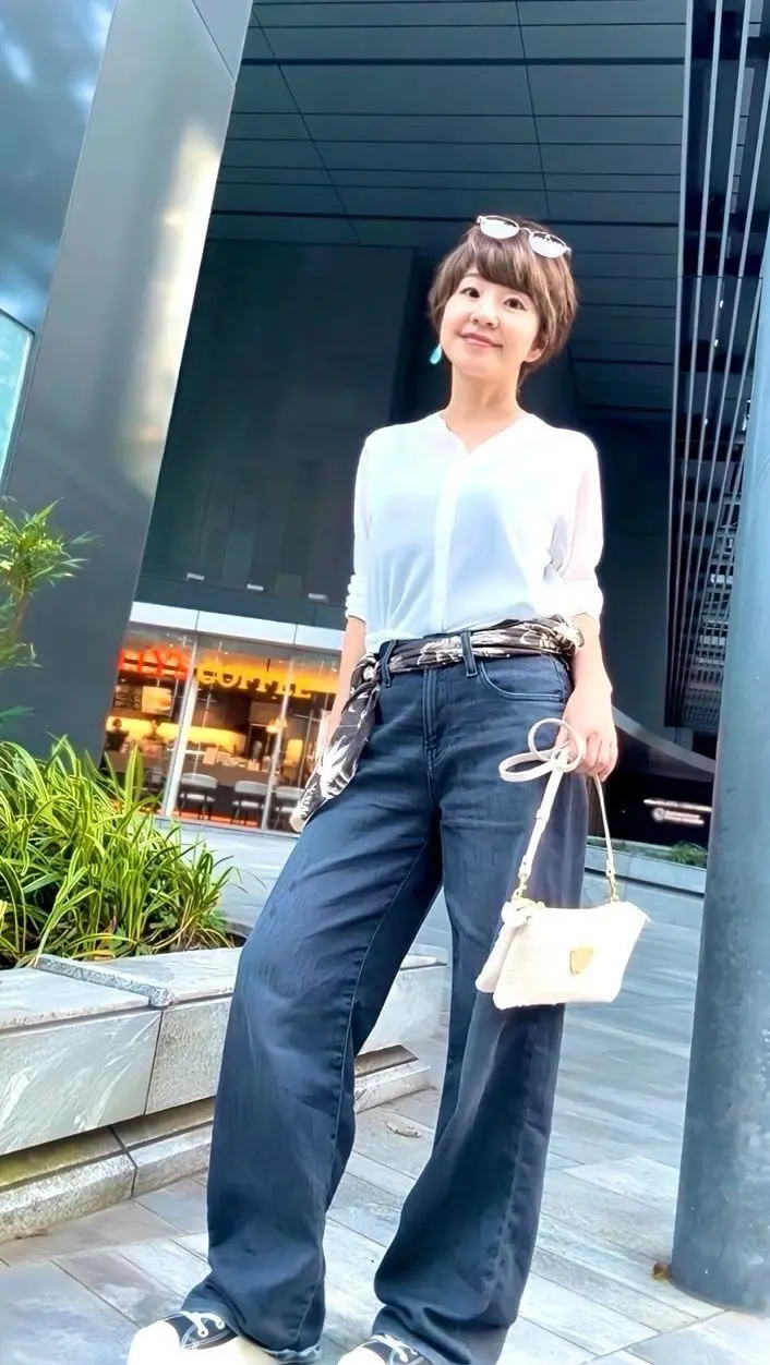 女性が白い長袖シャツとワイドパンツを着用し、屋外でポーズしています。頭頂にサングラスをかけ、右手に白いショルダーバッグを持ち、左手はポケットに挿しています。背景にはモダンな建物と緑の植物が見えます。