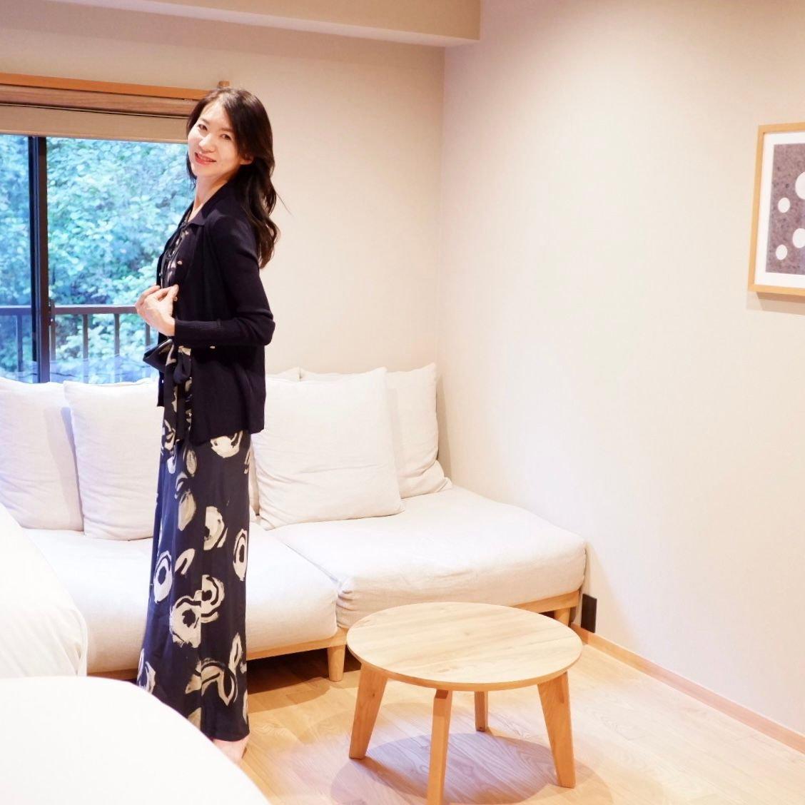 明るいリビングで、黒いカーディガンと花柄のロングワンピースを着た女性が立っている。窓から自然光が差し込み、白いソファと木製の丸テーブルが配置されている。壁には抽象的なアートが飾られている。