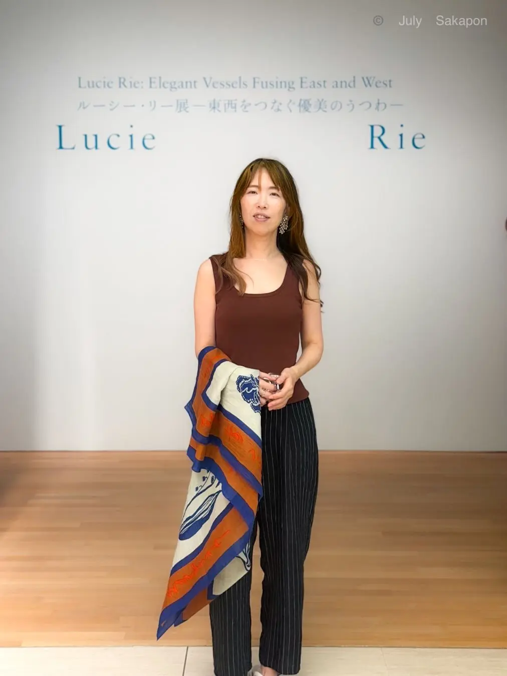展示会場で、女性が作品を手に立っている。茶色のタンクトップにストライプのパンツを着用し、カラフルな織物作品を持つ。背景には「Lucie Rie」と書かれた展示タイトル。
