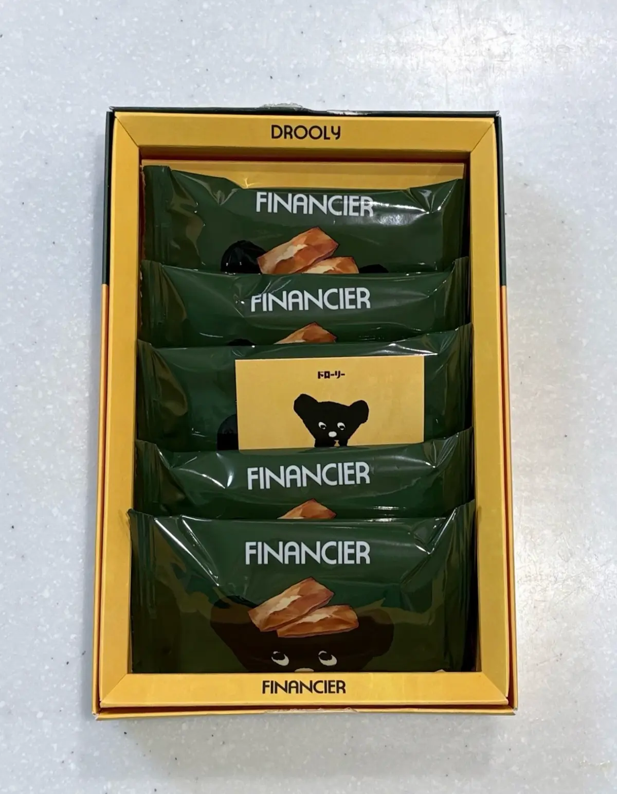 ゴールデン色の箱の中に、緑色の包装された「FINANCIER」のチョコレートが5個入っている。包装にはチョコレートの絵と小さなクマのキャラクターが描かれている。