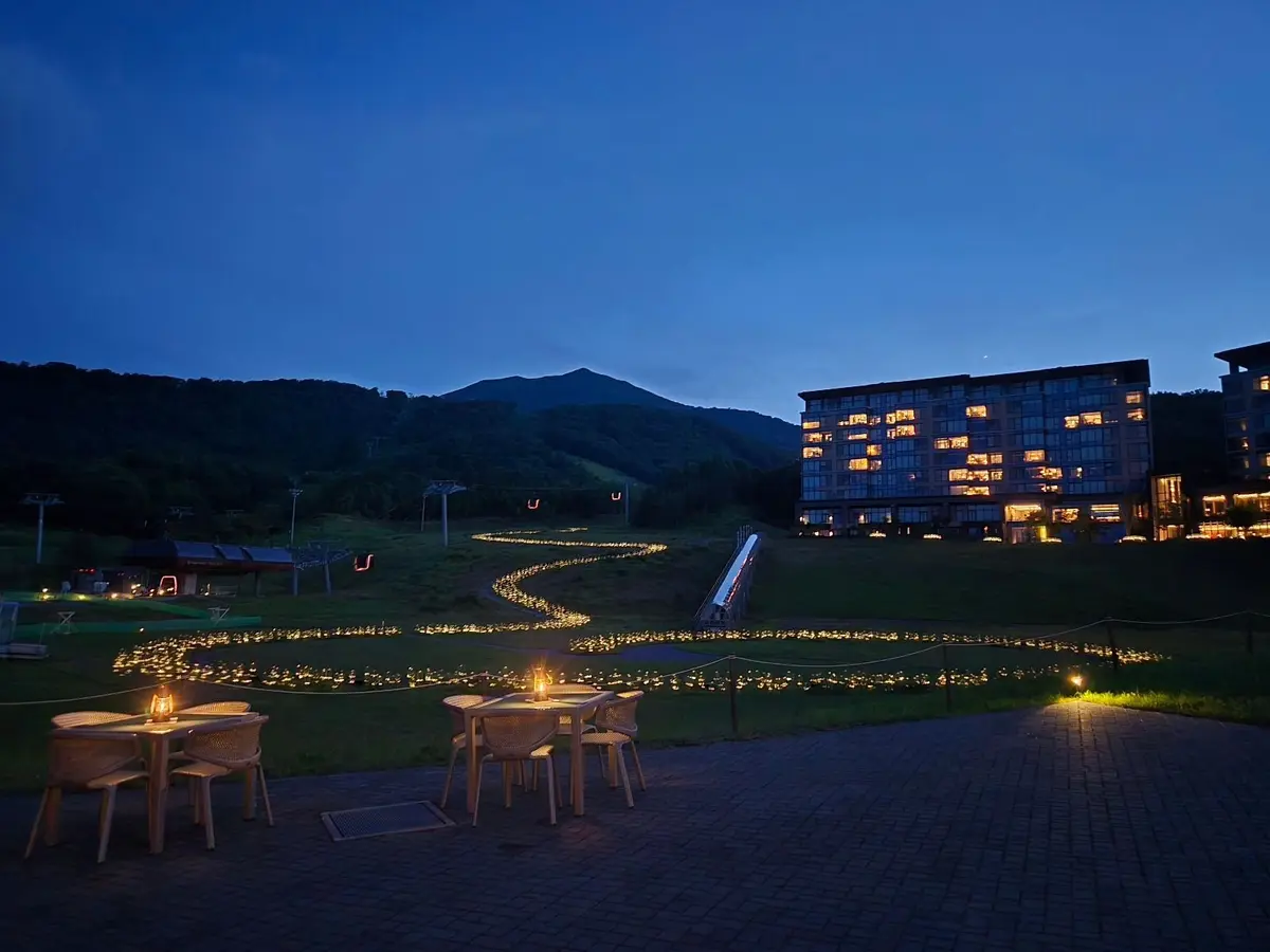 夜の青空を背景に、緑の芝生にテーブルと椅子が並び、小さな光が灯されている。遠景には、窓が光る大型のホテルと、山々が連なっている。