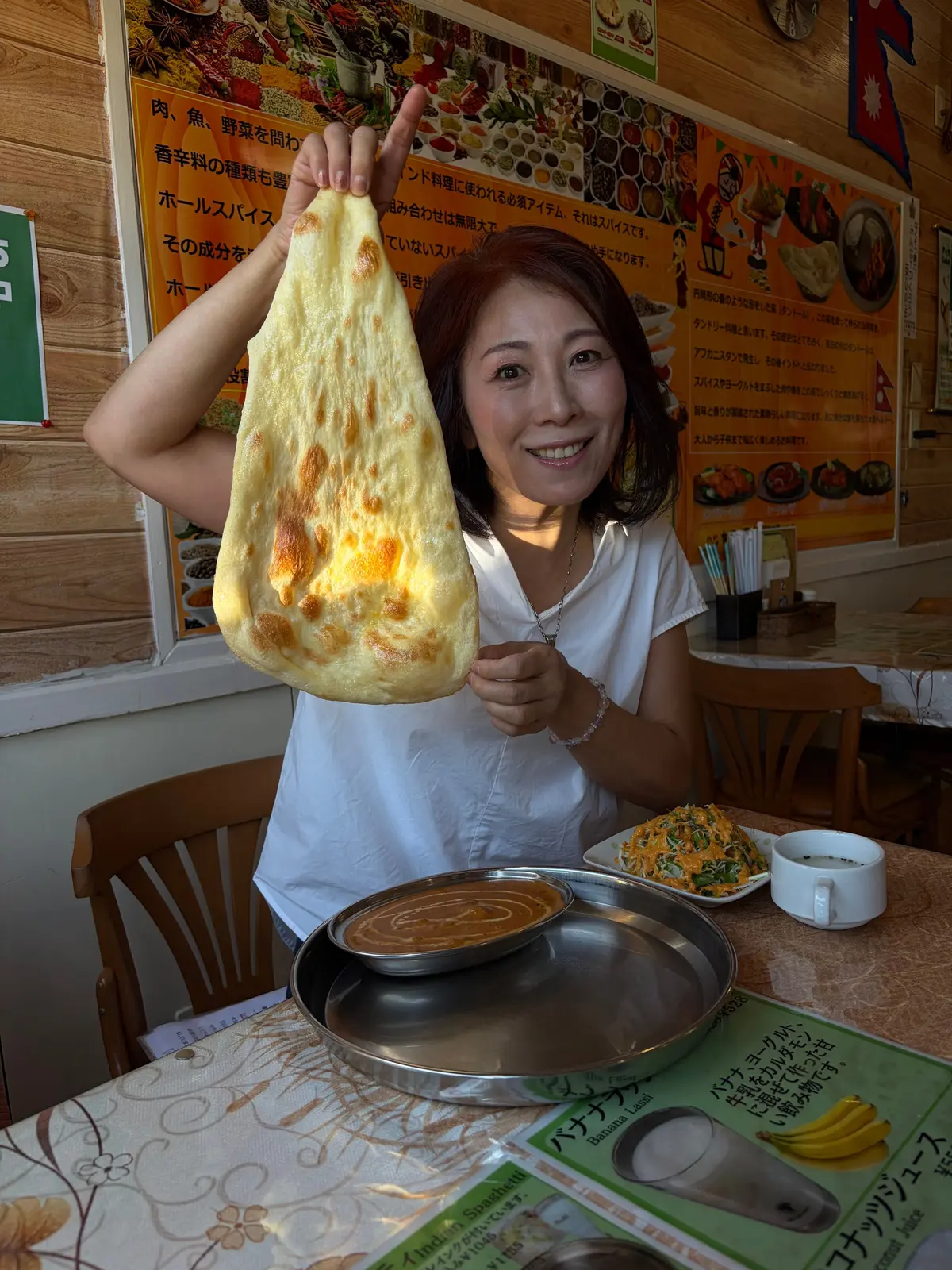 笑顔の女性が、金属製の皿に入ったカレーを巨大なドーサで包み、掲げている。白いシャツを着ており、背景には料理の写真やメニューが飾られている。