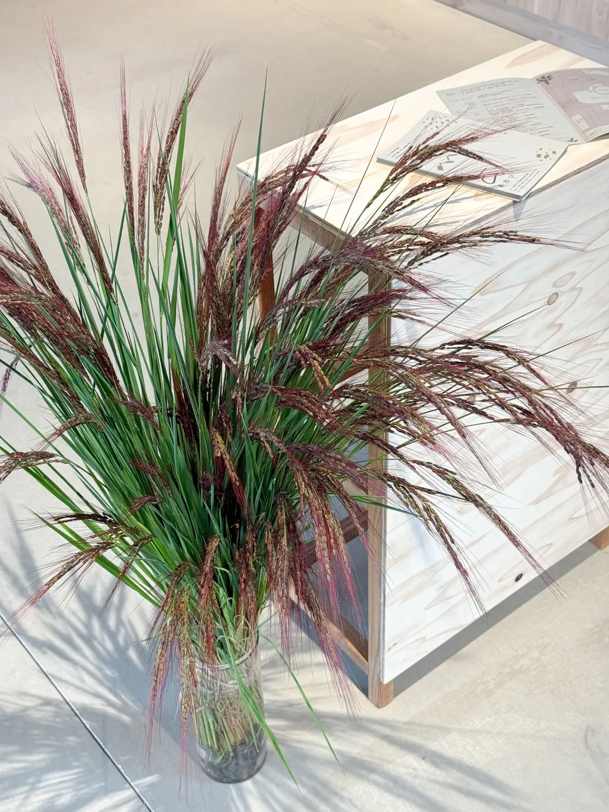 ガラス瓶に入った穂状の植物の装飾。緑と赤紫色の穂が密集し、木製のテーブルの横に置かれている。テーブルには書類が置かれている。