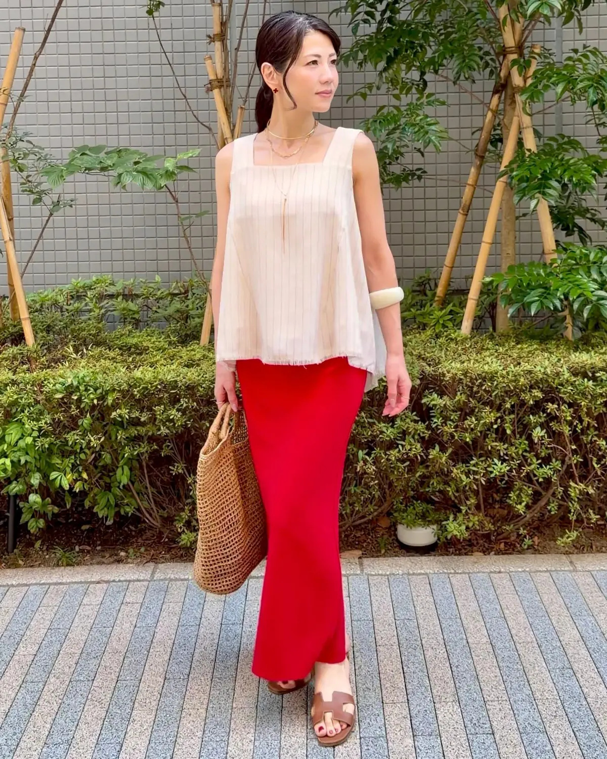 ベージュのトップスと赤色のフレアパンツを着用した女性が、屋外のタイル張りの道でポーズをとっている。右手に編み込みのバッグを持ち、ブラウンのサンダルを履いている。背景には緑の植栽とグレーの壁がある。
