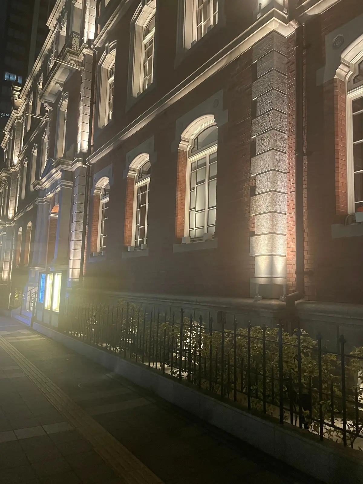 夜の街並み。赤レンガの建物がライトアップされ、アーチ型の窓が並ぶ。手すり沿いの緑地帯があり、歩道には点字ブロックが見える。霧気味の雰囲気。
