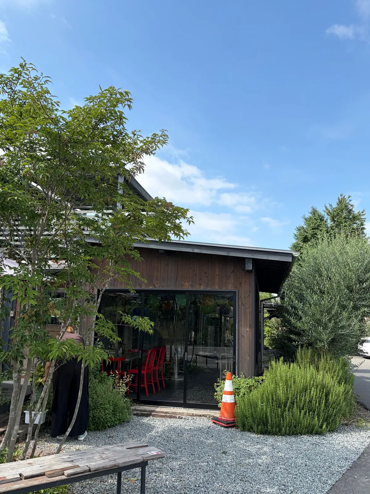 木造の建物と、赤色の椅子が並ぶテラス席。手前に木々が生い茂り、オレンジ色のコーンが置かれている。背景は青空に白い雲。