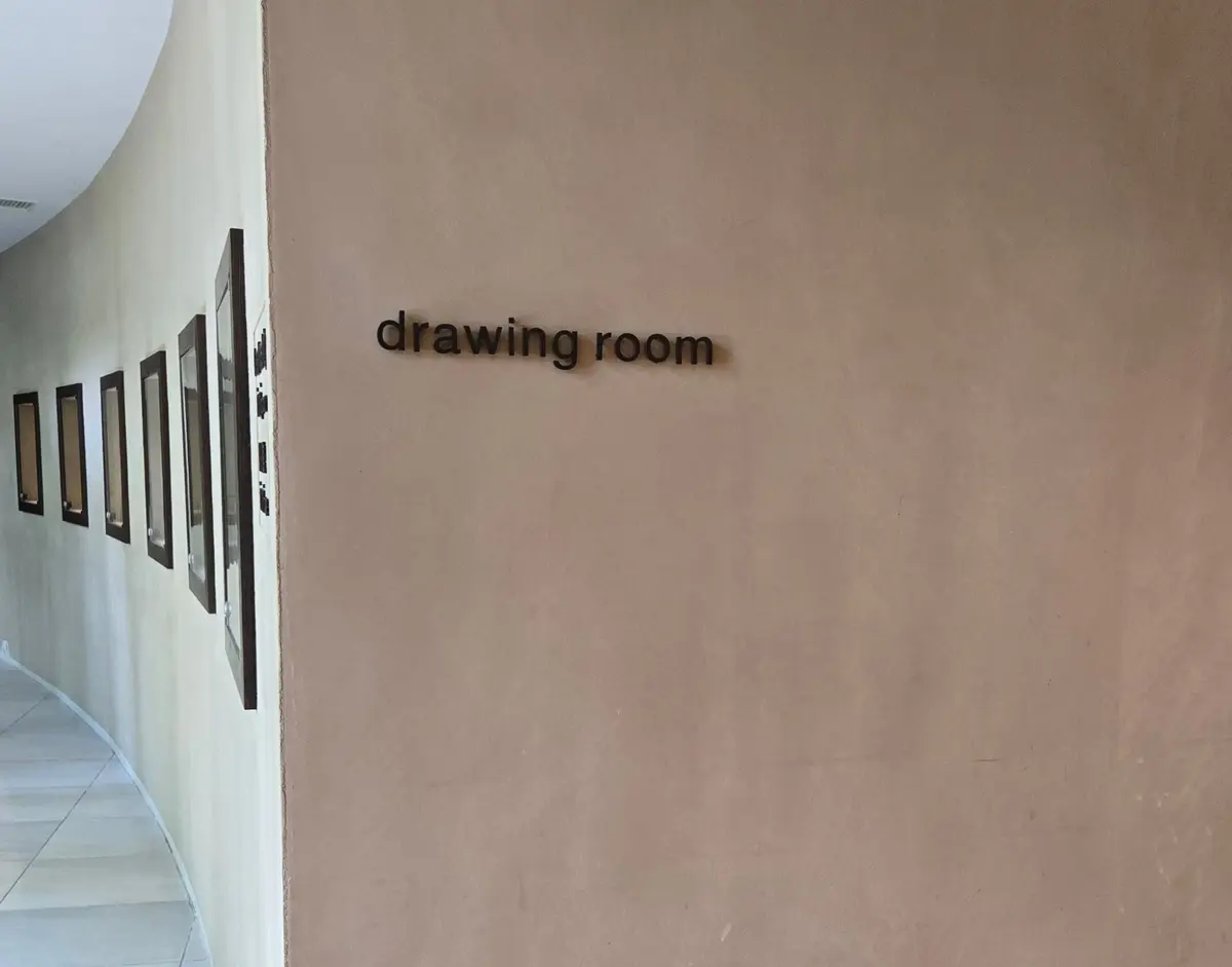 ベージュ色の壁に、黒色の文字で「drawing room（ドローイングルーム）」と表記されたサイン。左側には複数の額縁に入った絵画が並んでいる。