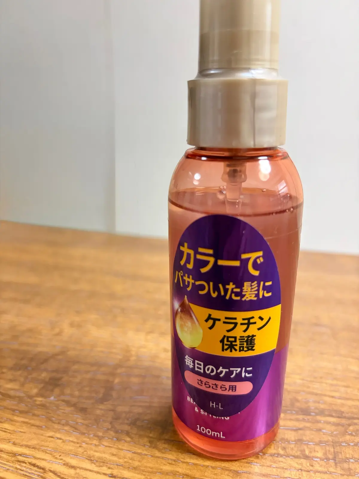 茶色のキャップが付いた、オレンジ色の液体が入ったスプレーボトル。正面には青と黄色の文字で「カラーでパサついた髪に」などの日本語のラベルが貼られている。100mlの容量表記あり。