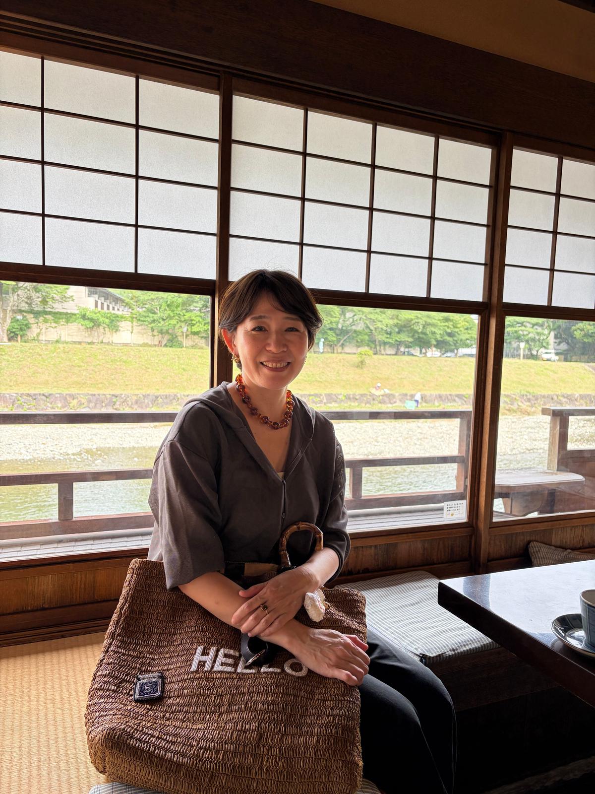 窓辺に座る女性。グレーのシャツを着て、オレンジ色のネックレスを身につけ、笑顔でこちらを見ている。大きな「HELLO」と書かれたバッグを持つ。背景には水辺と緑の風景が広がる。