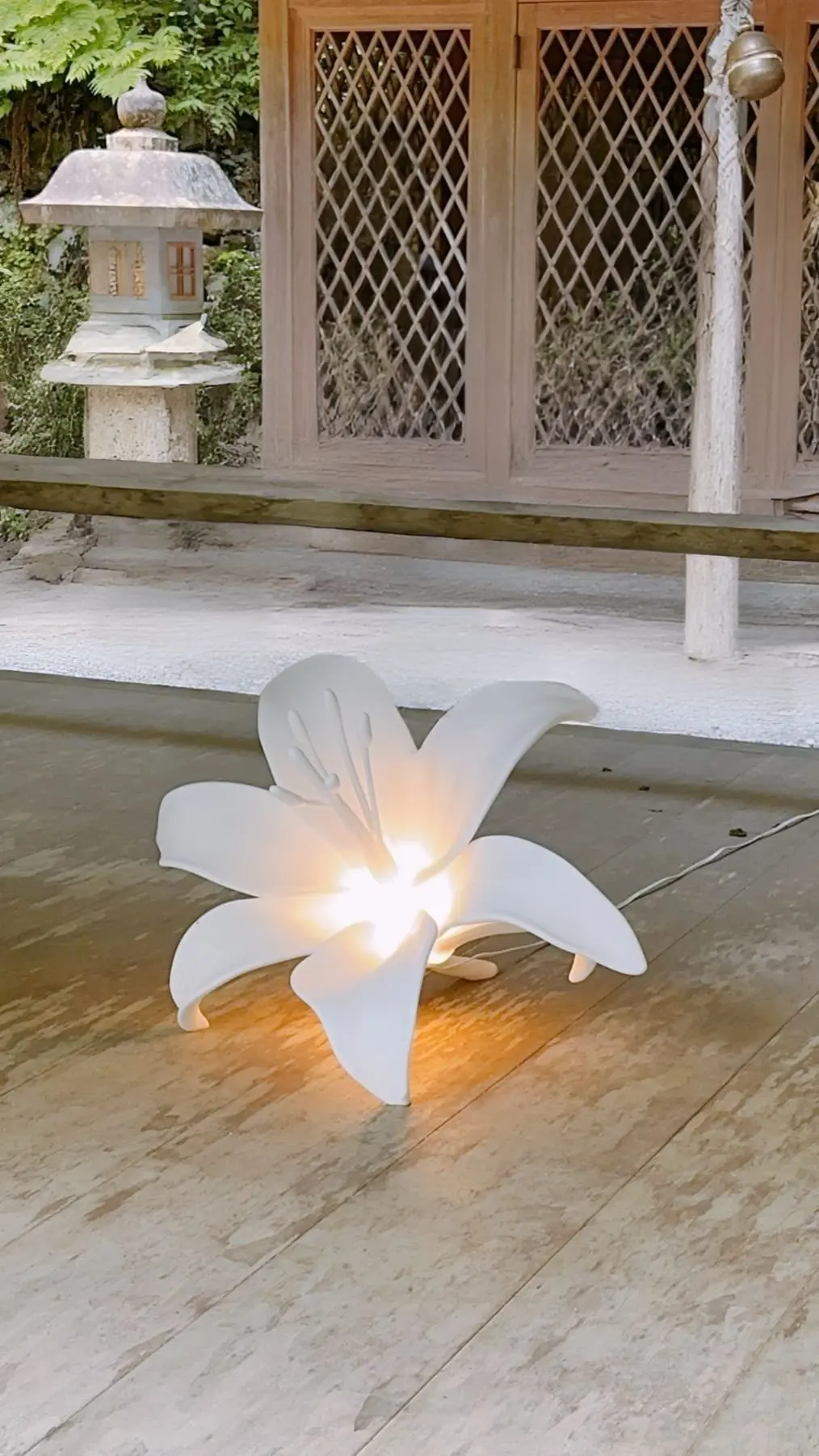 白い花の形をしたLEDランプが木製の床に置かれ、中心から光を放っている。背景には木製の建物と石の灯篭が見える。