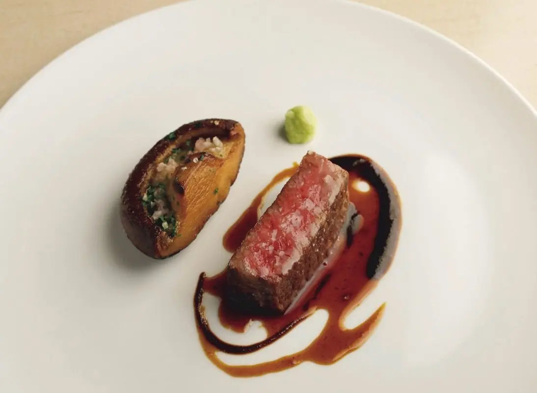 白い皿に盛り付けられた、赤身の肉と付け合わせ。肉は断面がピンク色で、照り気のあるソースが描かれている。隣にはきのこに具材が詰まったものと、緑色のペースト状のものが置かれている。