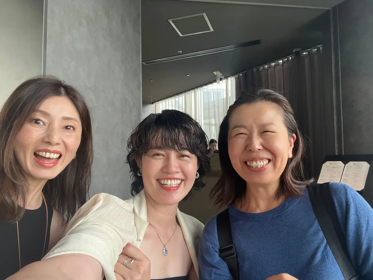 笑顔の3人の女性が写っているセルフィー写真。左から茶髪の女性、黒髪の女性、白髪混じりの女性。背景には窓とカーテンが見える。