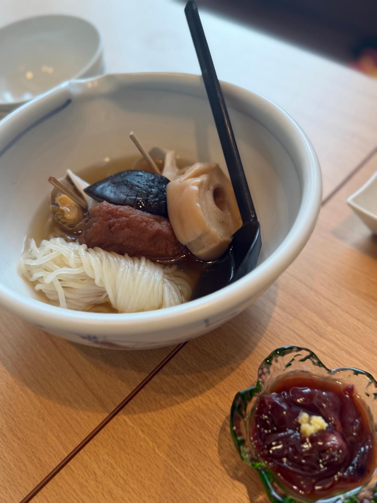 白い器に入った、白玉とキノコ、黒い練り物が入った汁物の料理。黒い箸が添えられ、右下には赤紫色のゼリー状のものがガラスの器に入っている。